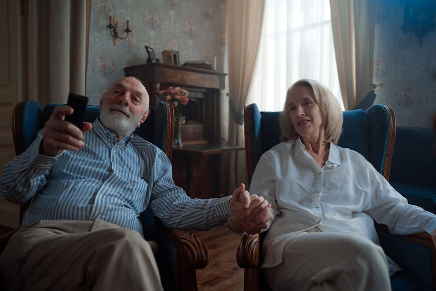Older man and woman in armchairs holding hands while watching television
