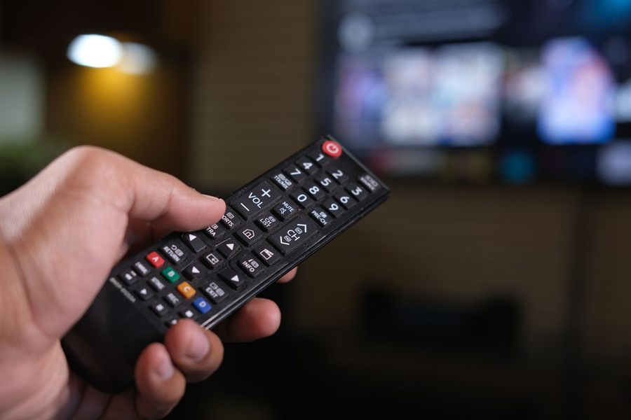 Close-up of a hand holding a TV remote with the screen glowing behind